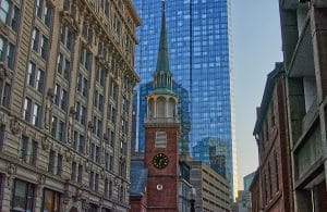 A historic brick church with a tall green steeple and clock stands between older stone buildings, with a modern glass skyscraper rising behind—an iconic view perfect for a School Trip to Boston, all bathed in daylight.