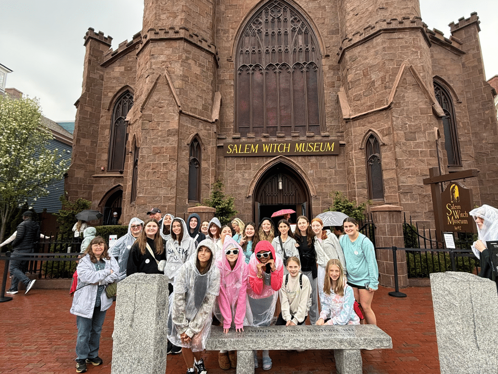A group of people wearing rain ponchos pose and smile in front of the Salem Witch Museum, a large brown stone building with arched windows and a sign above the entrance. It appears to be a rainy day.