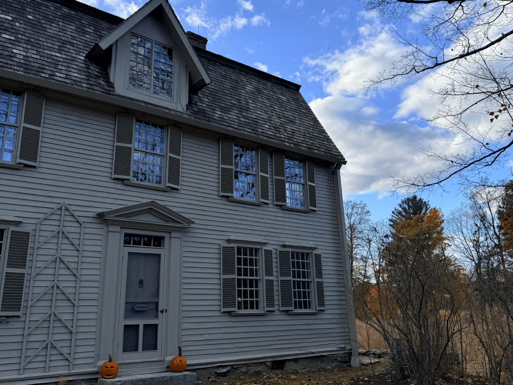 A gray wooden house with shuttered windows stands under a partly cloudy sky. Two carved pumpkins sit by the front door, and bare trees surround the house with autumn foliage visible in the background.