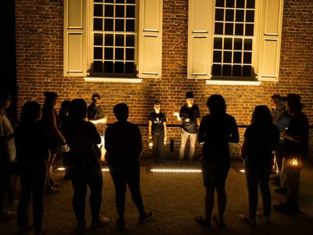 A group of people stand in a circle at night, illuminated by warm lights, in front of a brick building with two large windows and white shutters. Two individuals stand at the center, appearing to address the group.