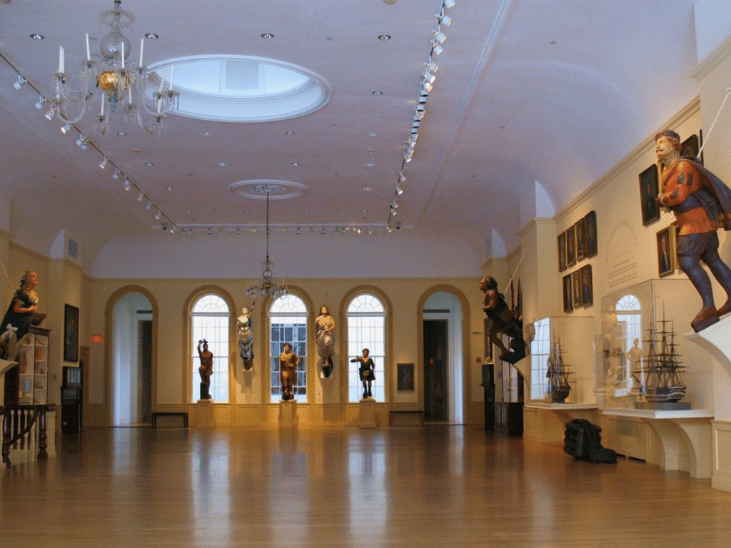 A spacious museum gallery with wooden floors, ship figureheads displayed along the walls, model ships in glass cases, large arched windows at the back, and elegant chandeliers hanging from the ceiling.