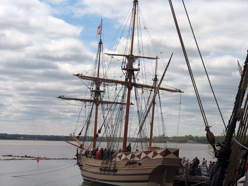 A replica of a historic wooden sailing ship with tall masts and rigging is docked at a pier under a cloudy sky. Several people, perhaps on a Colonial Williamsburg Field Trip, stand on the deck and dock by the river in the background.