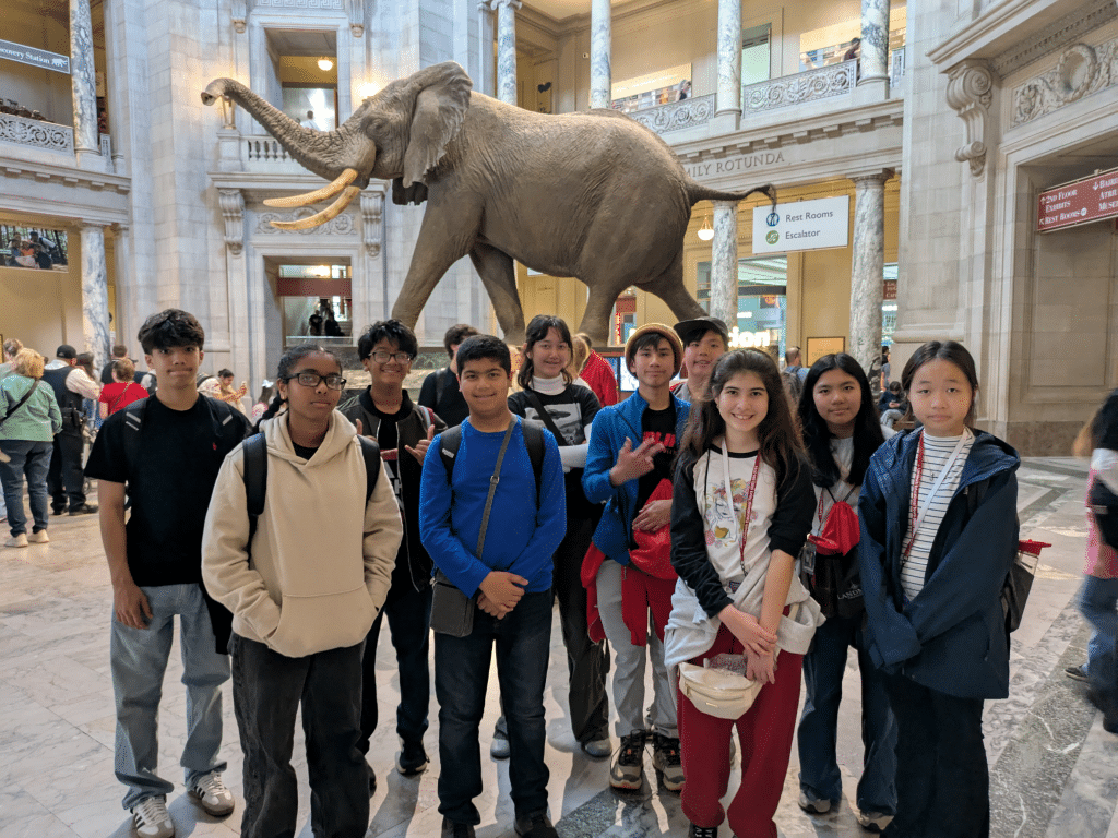 A group of ten kids poses for a photo inside a museum, standing in front of a large taxidermy elephant display. The museum has marble floors, columns, and balconies with people in the background.