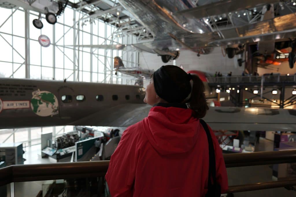A person in a red jacket looks up at vintage airplanes displayed in a spacious, well-lit aviation museum—one of the top STEM Washington DC school trip ideas—with large windows and metal railings.
