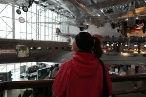 A person in a red jacket looks up at vintage airplanes displayed in a spacious, well-lit aviation museum—one of the top STEM Washington DC school trip ideas—with large windows and metal railings.