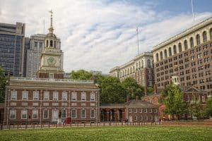 Independence Hall in Philadelphia, a historic red-brick building with a clock tower, stands amid trees and modern office buildings under a partly cloudy sky. People on a field trip to Philadelphia walk nearby, with a grassy area in the foreground.