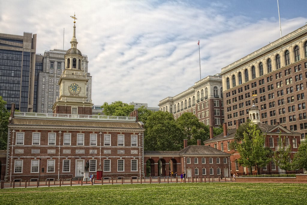 Independence Hall in Philadelphia, a historic red-brick building with a clock tower, stands amid trees and modern office buildings under a partly cloudy sky. People on a field trip to Philadelphia walk nearby, with a grassy area in the foreground.