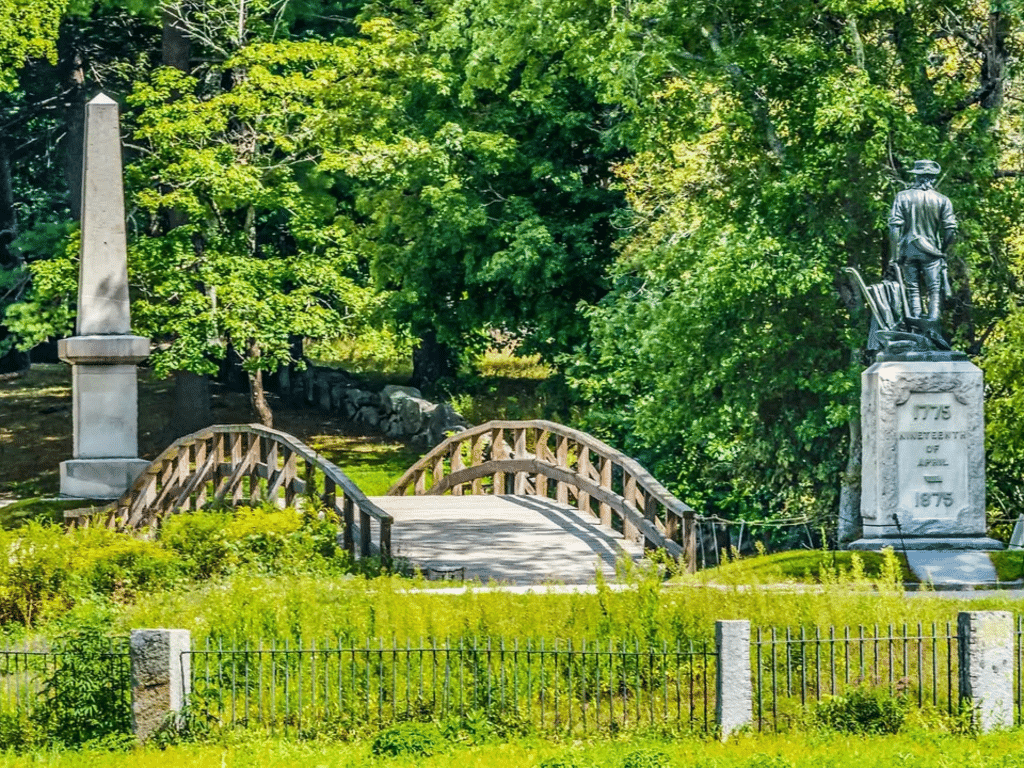 A small wooden bridge crosses a grassy area near a stone obelisk and a statue of a soldier on a pedestal, surrounded by lush green trees and bushes.