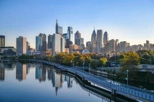 Philadelphia skyline with modern skyscrapers and a tree-lined riverwalk reflecting in the calm river under a clear blue sky, offering students stunning views while exploring Philadelphia historical sites for students nearby.