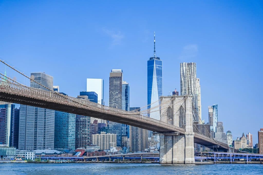 The Brooklyn Bridge spans across the water with the New York City skyline, including the One World Trade Center, visible in the background under a clear blue sky—a stunning highlight for Educational Student Tours in NYC.