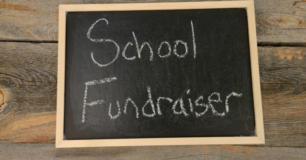 A handheld chalkboard sits on a wooden tabletop surface with chalk writing that says "school fundraiser."