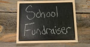 A handheld chalkboard sits on a wooden tabletop surface with chalk writing that says "school fundraiser."