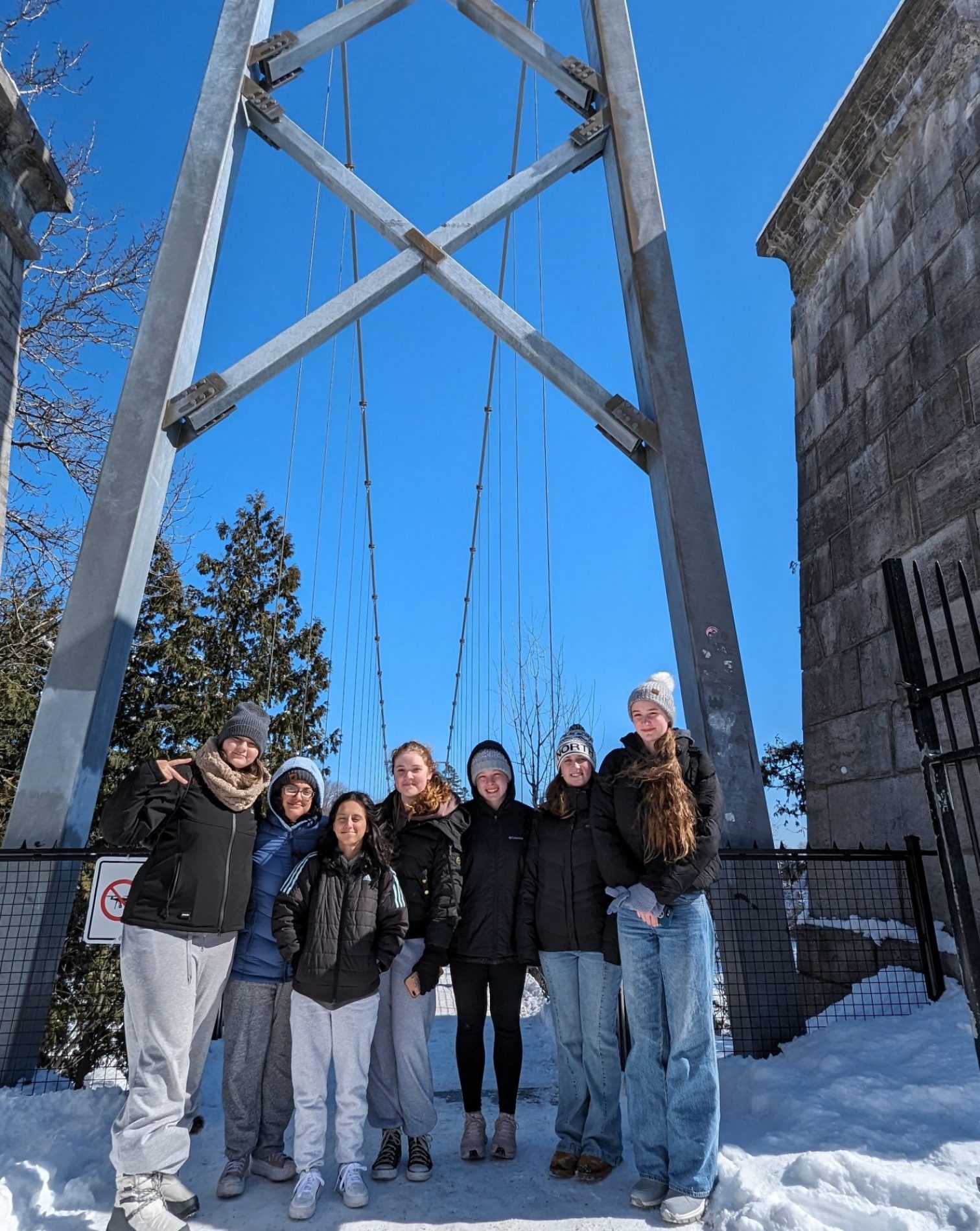 A group of seven people bundled in winter clothing stands on a snowy suspension bridge. The sky is clear and blue, and the bridge's metal structure is visible above them. Trees and part of a stone building frame the scene.